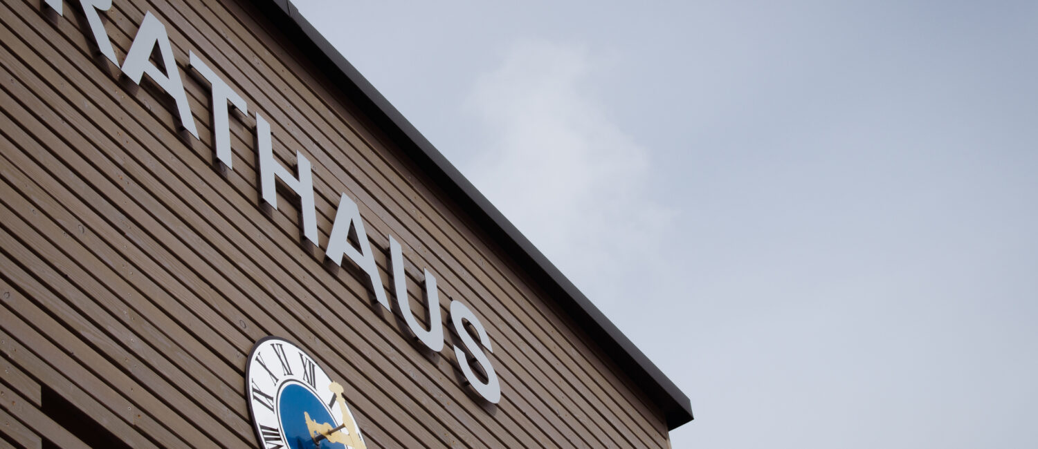Frontseite des Rathauses Parkstetten, Aussenfassade mit braunen Holz verkleidet, darauf eine große Uhr und darüber ist mit großen silbernen Buchstaben Rathaus zu lesen.
